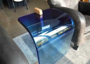 A transparent blue resin waterfall tabletop with a single wooden leg sits on a grey rug between two grey leather chairs.